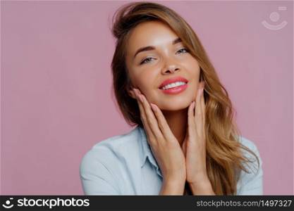 Photo of lovely young female model touches gently chin with both hands, smiles gently, has tender look, long hair combed on right side, wears professional makeup, isolated over purple background