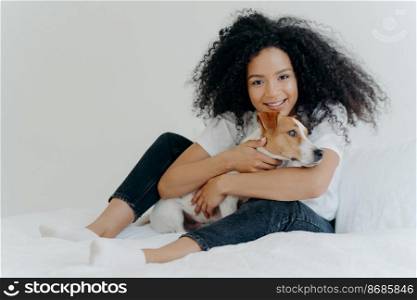 Photo of lovely young female pet owner poses in clean white bedroom, hugs dog, plays with best friend, wears casual clothing, has cheerful expression. People, animals, love, friendship concept