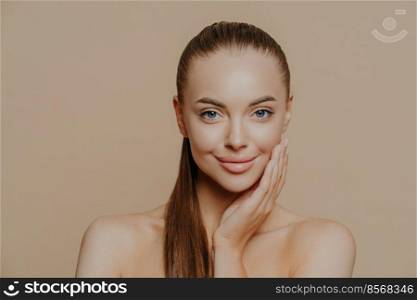 Photo of lovely young woman has smooth healthy skin after beauty procedures and receiving facial massage, stands bare shoulders, has long dark straight hair, isolated over beige wall cares of her body