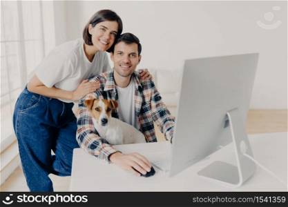 Photo of male freelancer works remotely at computer with dog, his affectionate wife embraces and helps with some questions, pose in big room at workplace, discuss some issues and family budget