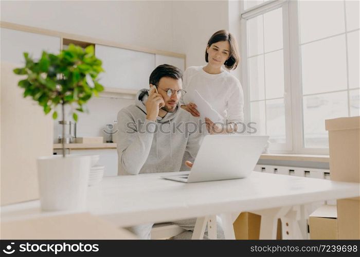 Photo of male model searches information on laptop computer, searches new flat for rent, calls client via mobile phone, woman stands near with paper documents, check domestic bills, pose in kitchen