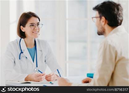 Photo of male patient writes all details makes notes during consultancy with doctor sit in spacious office. Physician tells treatment issues and explains diagnosis to hospital visitor. Medical concept