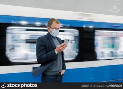 Photo of man in eyewear and protective medical mask, uses modern phone, travels by public transport, stands against train in motion on platform, gets information online about new Covid-19 virus