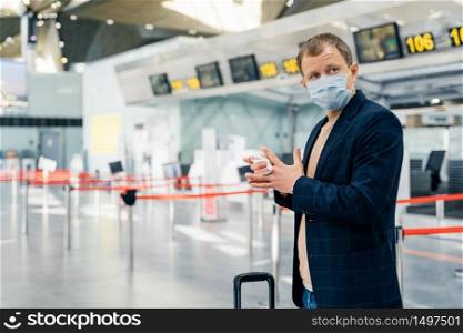 Photo of man passenger poses in airport, wears medical mask, uses antiseptic napkin to prevent spreading bacteria and coronavirus, cares about antibacterial treatment in public crowded place