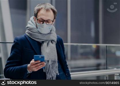 Photo of man watches news on his phone, wears protective mask, protects from air contamination or coronavirus, checks email box, wears warm scarf. Covid-19. Epidemic disease. Health, social media