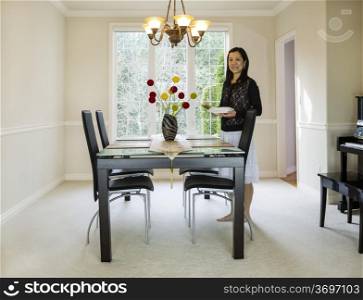 Photo of mature woman placing diner plates and chop sticks in family formal dining room with daylight coming through large windows in background