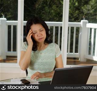 Photo of mature woman, showing stress, working at home with laptop, cell phone and papers on top of table and large windows in background