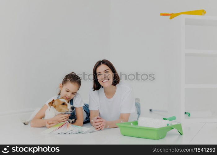 Photo of mother, daughter and pedigree dog lie on floor, take break after painting walls, surrounded with tray and paint roller, isolated over white background, busy with housework and repair