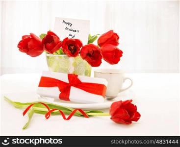 Photo of of beautiful tulips pot with greeting postcard, gift box and cup of coffee on the table at home, spring season, happy mothers day, still life, morning beverage in holiday for mom