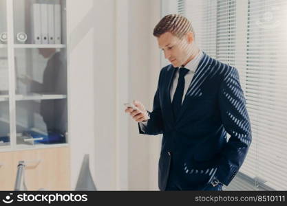 Photo of office employee in elegant outfit, has appealing appearance, uses modern cell phone, checks notification, poses in spacious cabinet, connected to wireless internet. Networking concept