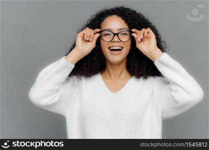 Photo of optimistic lovely woman looks happily through spectacles, keeps hands on rim of spectacles, notices something pleasant, wears white sweater, isolated over grey background. Happiness