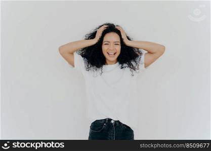 Photo of overjoyed curly haired woman laughs hapπly, has fun, dressed in white t shirt and jeans, smi≤s broadly, isolated over white background. Peop≤, emotions, hapπ≠ss, ethnicity concept