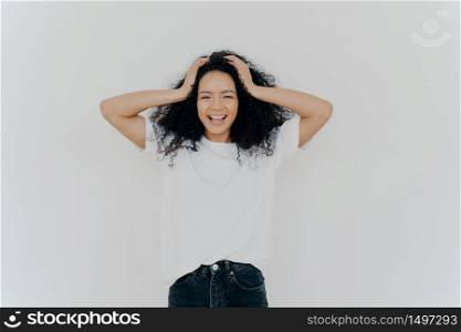 Photo of overjoyed curly haired woman laughs happily, has fun, dressed in white t shirt and jeans, smiles broadly, isolated over white background. People, emotions, happiness, ethnicity concept