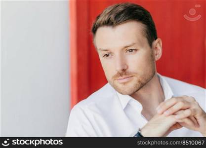 Photo of pensive male entrepreneur looks thoughtfully aside, keeps hands tgether, dressed in elegant white shirt, thinks about climbing on career ladder. People, business and thoughts concept