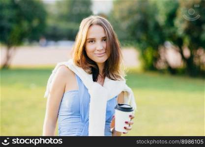 Photo of pleasant looking brunette young woman with pleasant appearance, spends weekend in open air, has stroll across park, holds paper disposable cup with beverage. People and leisure concept
