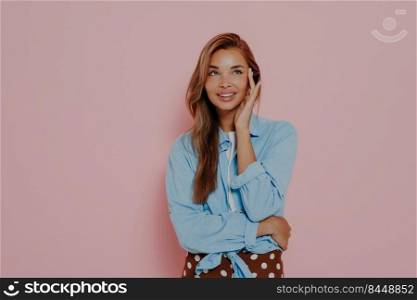 Photo of pleasant looking female model touches cheek with hand, has gentle lovely smile, wears light makeup, dressed in casual shirt and skirt, stands against pink wall, copy space for advertisement. Photo of pleasant looking female model touches cheek with hand
