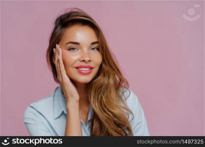 Photo of pleasant looking female model touches cheeks with palm, has gentle toothy smile, wears makeup, dressed in casual shirt, stands against purple wall, copy space area for your advertisement