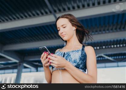 Photo of pleasant looking teenager with dark hair, holds modern cell phone, texts in social networks, listens favourite songs with earphones, wears casual top, stands outdoor. People and music concept