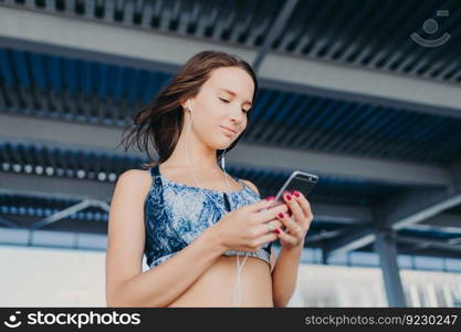 Photo of pleasant looking teenager with dark hair, holds modern cell phone, texts in social networks, listens favourite songs with earphones, wears casual top, stands outdoor. People and music concept