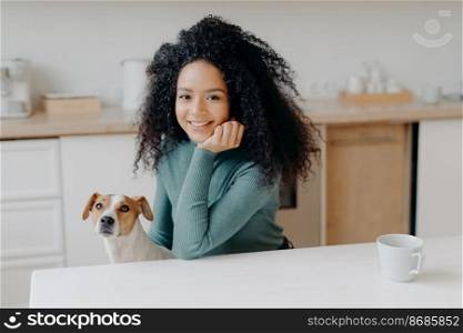 Photo of pleasant looking woman dressed in casual wear, has curly bushy hair, poses against kitchen interior with pedigree dog, has morning coffee, enjoys weekend, smiles pleasantly at camera