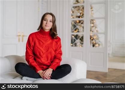 Photo of pleasant looking young woman dressed in red sweater, trousers, sits crossed legs on comfortable couch, enjoys domestic atmosphere, decorated New Year tree in background. Home concept