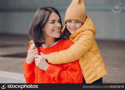 Photo of pleased attractive young mother looks positively at daughter, embraces mother, enjoy togetherness, have toothy smiles, eyes full of happiness, have good relationships. Family portrait