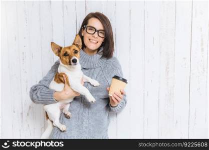 Photo of pleased brunette female carries her favourite dog, drinks hot coffee from paper cup, has pleasant warm smile on face, rejoices nice time spent with pet, poses against white fence background