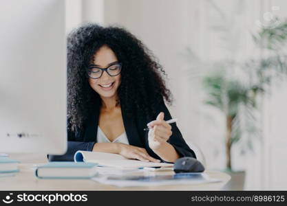 Photo of pleased curly haired woman writes down some information, holds pen, has smile, wears optical glasses and formal clothes poses at workplace with computer. Student writes ideas for course paper