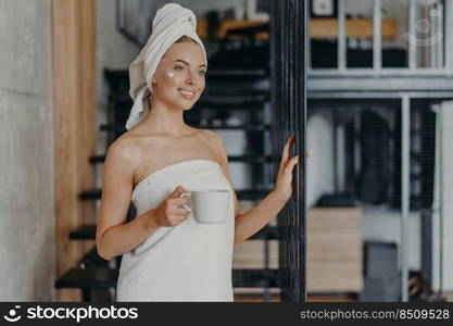 Photo of pleased smiling European woman enjoys day off at home, applies face cream, drinks tea, wrapped in towel, poses near strairs, thinks about something pleasant, has healthy glowing skin