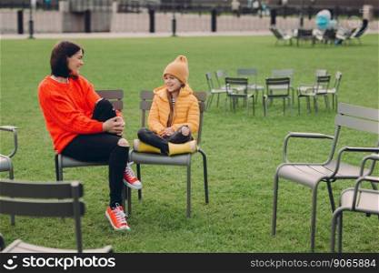 Photo of positive friendly young mother and daughter pose outside in recreation zone with green lawn on chairs, dressed in fashionable outfit, giggle at something funny. Family and communication