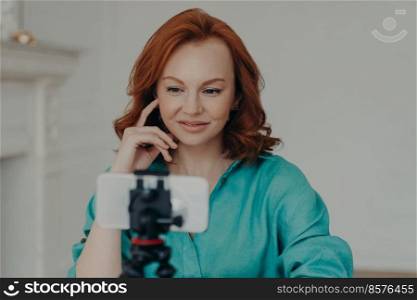 Photo of positive redhead woman blogger influencer shoots video for personal blog, uses modern mobile phone on tripod, does online chat, shoots blog at home, uses high speed internet connection
