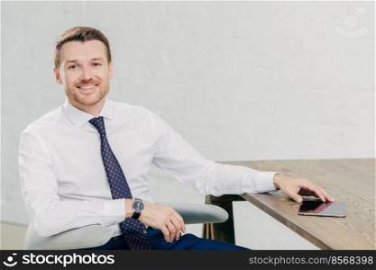 Photo of positve young successful male entrepreneur dressed in elegant white shirt with tie, looks positively at camera, has break after work, poses in his cabinet, uses modern tablet computer