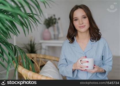 Photo of pretty female with appealing look, dressed in casual domestic clothes, holds white mug with aromatic coffee or tea, sits on wicker furniture indoor with copy space for your advertisement