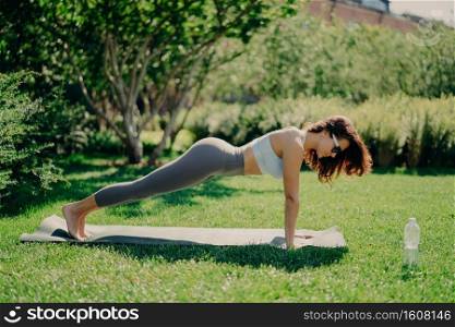 Photo of pretty fit woman poses full length in plank pose dressed in cropped top and leggings poses bare feet on fitness mat trains outdoors demonstrates endurance and motivation. Sport weight loss