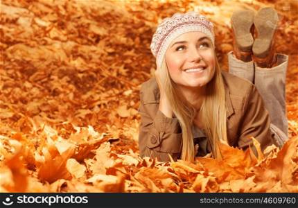 Photo of pretty girl lying down on the ground covered dry autumnal foliage, sweet blond girl wearing stylish warm hat, enjoying last autumn days, beautiful fall season, spending time in park
