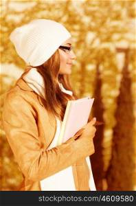 Photo of pretty student girl standing in autumn park and wearing beige leather coat with white warm hat, cute schoolgirl holding book, side view of beautiful teenager, back to school