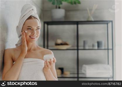 Photo of pretty young European woman applies moisturizing cream on face, has happy expression, wrapped in bath towel, stands against bathroom background indoor. Skin care and hygie≠concept
