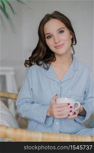 Photo of pretty young female looks directly at camera, dressed in stylish clothes, holds white mug with aromatic beverage, poses indoor at home, has good rest and pleasant talk with husband.