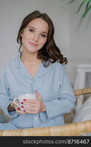 Photo of pretty young female looks directly at camera, dressed in stylish clothes, holds white mug with aromatic beverage, poses indoor at home, has good rest and pleasant talk with husband.