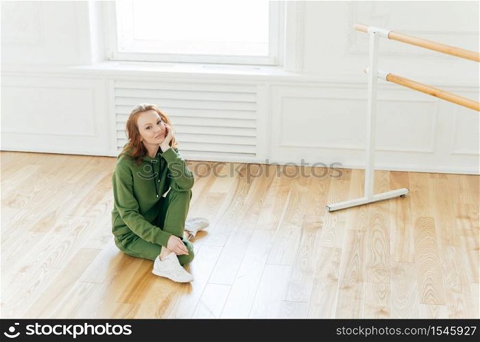Photo of professional female dancer has rest after training, poses on floor near ballet barre, has ballet classes, wears green sweatsuit, sportshoes, likes being active. People, fitness and exercising