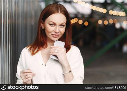 Photo of prosperous female business worker has break after work, sends text messages, holds cell phone in hand, dressed in white shirt, has browh hair, stands outdoor against blurred background