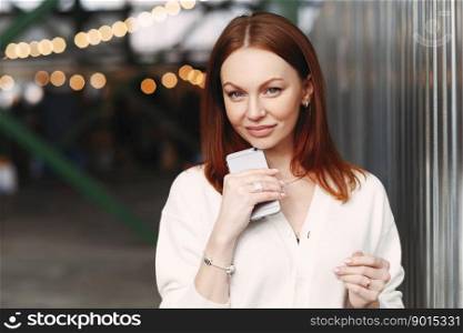 Photo of prosperous female business worker has break after work, sends text messages, holds cell phone in hand, dressed in white shirt, has browh hair, stands outdoor against blurred background