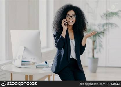 Photo of puzzled prosperous businesswoman calls partner, raises palm, holds mobile phone, wears spectacles and formal black outfit, tries to solve problem at work, discusses something actively