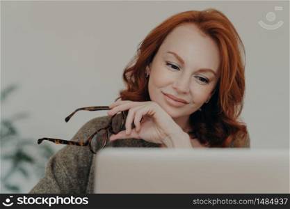 Photo of redhead woman takes off glasses, enjoys free time for e learning, focused at monitor of laptop computer, does distance job. Female student prepares for exams, uses modern technologies