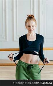 Photo of redhead woman with bun, wears black top, trousers, shows bare belly, has perfect body shape, has rehearsal, stands near ballet barre, conducts fitness lessons, being healthy and fit