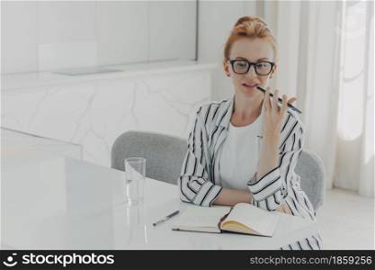 Photo of redhead young woman records voice call via smartphone microphone sits at desktop makes notes in notepad drinks fresh water uses voice search personal help service app records reminder. Photo of redhead young woman records voice call via smartphone microphone sits at desktop