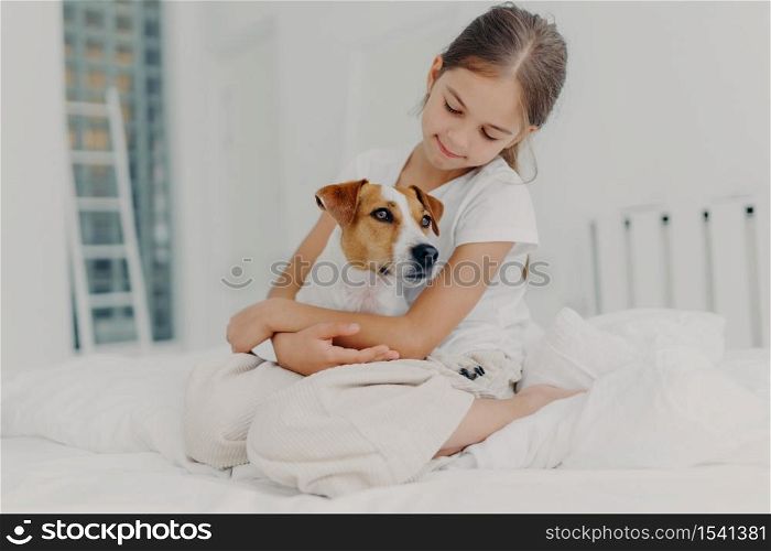 Photo of relaxed beautiful small girl plays with pedigree dog, embraces favourite animal, dressed in casual pyjamas, sits on bed, expresses love to pet, poses in light bedroom. Happy childhood concept