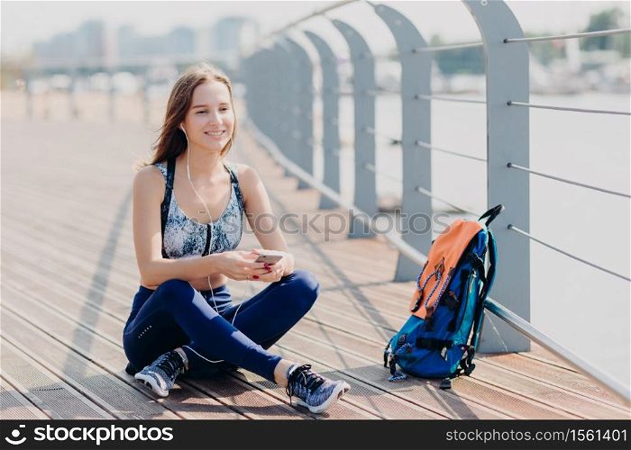 Photo of relaxed cheerful fitness girl in sporswear, sits crossd legs outdoor, rests after long walk, listens music with cellular and headphones, rucksack near. People, technology and recreation