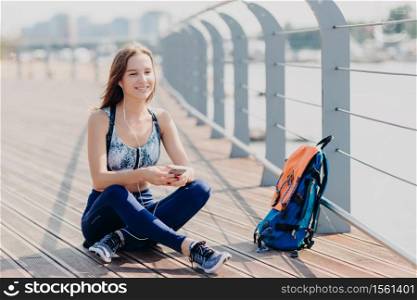Photo of relaxed cheerful fitness girl in sporswear, sits crossd legs outdoor, rests after long walk, listens music with cellular and headphones, rucksack near. People, technology and recreation