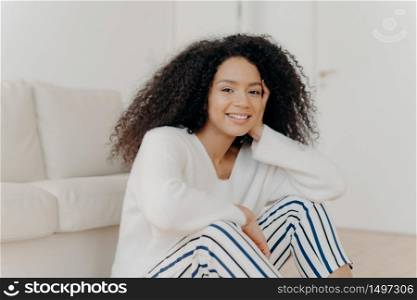 Photo of relaxed delighted African American woman with curly hairstyle, smiles gently at camera, wears white sweater and striped trousers, sits on floor near couch in living room of modern apartment